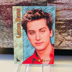 May include: A trading card featuring Lance Bass with the text "Lance Bass" in orange and yellow. The card has a light blue background and a red shirt. The *NSYNC logo is visible.