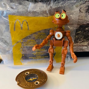 Puede incluir: Un juguete de plástico marrón con forma de robot con extremidades articuladas y ojos verdes, junto a una brújula circular. El robot tiene un emblema blanco en el pecho. El embalaje tiene el texto "TREASURE PLANET".