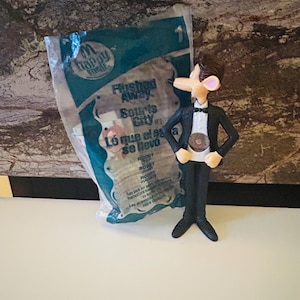 May include: A plastic toy figure of Roddy St. James from the animated film "Flushed Away," wearing a black tuxedo and bow tie. The figure is standing next to a Happy Meal bag with text in English and Spanish.
