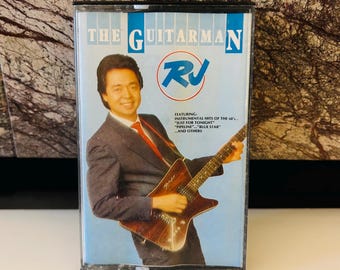 May include: A cassette tape with a blue background and a picture of a man playing a guitar. The text on the cassette tape reads "The Guitarman RJ Featuring: Instrumental Hits of the 60's "Just for Tonight" "Pipeline" "Blue Star" and others."