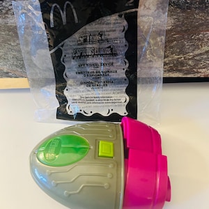May include: A gray and pink toy spy signal device with a green button and a clear plastic bag with text. The toy has a circuit board design and the text "SPY KIDS" is visible.