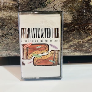 Puede incluir: Un casete con el álbum "Ferrante & Teicher: A Few of Our Favorites On Stage". La portada muestra dos pianos estilizados en tonos naranja y marrón, con títulos de canciones como "Memory" y "St. Louis Blues".