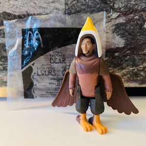 Könnte beinhalten: Brother Bear-Actionfigur von McDonald's, mit einer Figur mit gelb-weißem Kopfschmuck, braunen Flügeln und orangefarbenen Füßen. Die Figur befindet sich in einer transparenten Plastiktüte mit dem McDonald's-Logo.