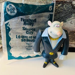 May include: A Whitey toy figure from the animated film "Flushed Away" wearing a black suit with a gold chain and a white collar. The figure is standing in front of a Happy Meal bag with text in English, French, and Spanish.