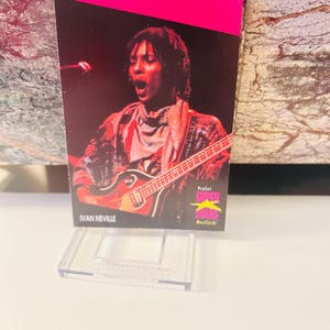 May include: A ProSet Super Stars Musicards trading card featuring Ivan Neville playing a guitar. The card has a pink border and the text "Ivan Neville" is printed at the bottom. The card is displayed in a clear plastic holder.