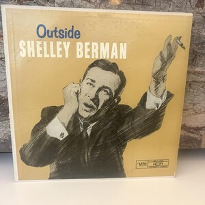 Può includere: Una copertina di un disco in vinile vintage con un'immagine in bianco e nero di un uomo in abito, che tiene una sigaretta. La copertina è gialla e presenta il testo "Outside" e "Shelley Berman".