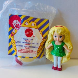 May include: A yellow plastic doll with long blonde hair wearing a green and white polka dot dress and red shoes. The doll is packaged in a clear plastic bag with a yellow and purple cardboard backing that says "Mattel" and "Under-3 Toy".