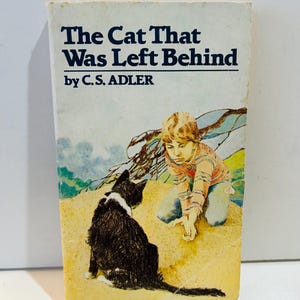 Könnte beinhalten: Ein Vintage-Taschenbuch mit dem Titel "The Cat That Was Left Behind" von C.S. Adler. Die Illustration auf dem Cover zeigt eine schwarz-weiße Katze und ein Kind. Das Kind kniet auf sandigem Untergrund, mit blau-grünem Hintergrund.