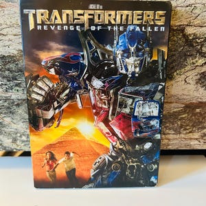 Op de afbeelding: DVD van de film "Transformers: Revenge of the Fallen." De cover toont Optimus Prime in rood, blauw en zilver, met de titel van de film bovenaan. Twee mensen rennen voor een piramide, op een donkere achtergrond.