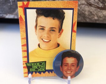 May include: A yellow and red trading card with a photo of a young man with dark hair. The card has the text "New Kids on the Block" on it. A blue button with a photo of the same young man is next to the card.