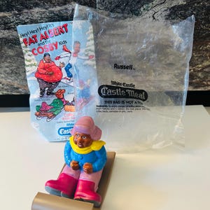 May include: A vintage Fat Albert and the Cosby Kids toy figure on a sled. The figure wears a blue jacket, pink boots, and a pink hat. The packaging includes a White Castle Castle Meal bag.