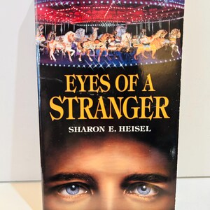 May include: A paperback book titled "Eyes of a Stranger" by Sharon E. Heisel. The cover features a carousel scene with horses and a night sky. Below the title are a pair of blue eyes.