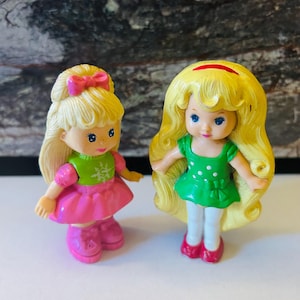 May include: Two small plastic dolls with long blonde hair. One doll is wearing a pink dress with a green sweater and a snowflake design. The other doll is wearing a green dress with white polka dots and a white bow.