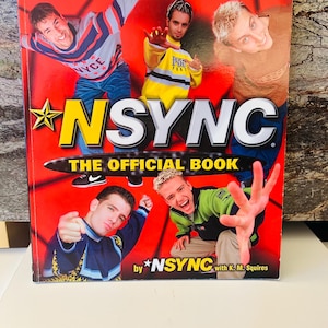 Op de afbeelding: Een rode boekomslag met de band *NSYNC en de tekst "NSYNC THE OFFICIAL BOOK" in geel en zwart. De omslag heeft afbeeldingen van de bandleden. Het boek is van *NSYNC met K. M. Squires.