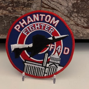 Könnte beinhalten: Ein runder, blauer und roter gestickter Aufnäher mit den Worten "PHANTOM FIGHTER F4D" in Weiß. Der Aufnäher zeigt die Silhouette eines Düsenflugzeugs, das über einer Stadtlandschaft fliegt.