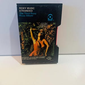 May include: A black 8-track tape of the Roxy Music album "Stranded." The album cover features a woman in an orange dress. The text "ROXY MUSIC STRANDED" is visible on the tape.