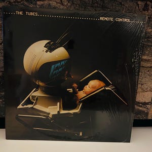 Könnte beinhalten: Ein schwarzes Vinyl-Plattencover mit einem Retro-Fernseher, in dem sich ein Baby befindet. Das Albumcover trägt den Text "THE TUBES" und "REMOTE CONTROL".