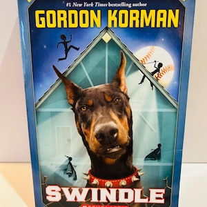 May include: A hardcover book titled "Swindle" by Gordon Korman. The cover features a Doberman Pinscher wearing a spiked collar, with a baseball and silhouettes of people. The book is published by Scholastic and is a #1 New York Times bestseller.