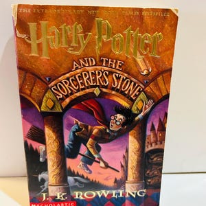 May include: A hardcover book titled "Harry Potter and the Sorcerer's Stone" by J.K. Rowling. The cover features a vibrant illustration of Harry Potter on a broomstick, with a castle and columns in the background. The Scholastic logo is visible.