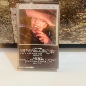 May include: A vintage cassette tape of The Cars album. The cover features a close-up of a person's face. The tracklist includes songs like "Good Times Roll" and "Bye Bye". The cassette is in a clear plastic case.