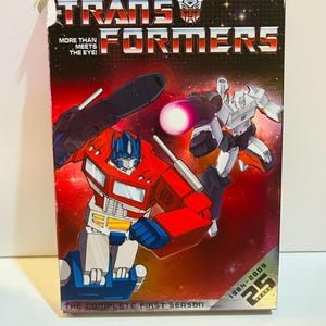 Op de afbeelding: Een Transformers The Complete First Season dvd-boxset. De doos toont Optimus Prime en Megatron in actieposities tegen een rode en zwarte ruimteachtergrond. Tekst bevat "More Than Meets The Eye!" en "25 Years 1984-2009".