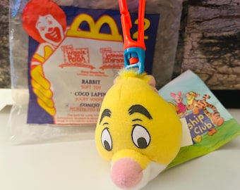Könnte beinhalten: Ein gelbes Plüschtier, das Rabbit darstellt, mit einer rosa Nase und schwarzen Augenbrauen, befestigt an einem roten und blauen Clip. Das Spielzeug stammt aus der Winnie-Puuh-Serie und befindet sich noch in der Originalverpackung.