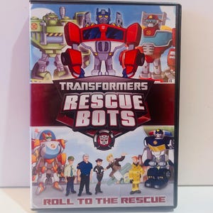 Op de afbeelding: DVD-hoesje met de Transformers Rescue Bots. De cover toont verschillende robotkarakters in rood, groen en blauw, met de titel "Transformers Rescue Bots" prominent weergegeven. De zin "Roll to the Rescue" is ook zichtbaar.