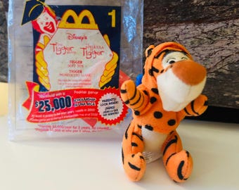 Könnte beinhalten: Ein kleines, plüschiges Tigger-Spielzeug aus Disneys Tigger-Film, in einer durchsichtigen McDonald's Happy Meal-Tüte. Das Spielzeug ist orange mit schwarzen Streifen und Punkten.