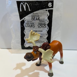 Könnte beinhalten: Eine braune und orangefarbene Elchfigur mit großen Geweihen, Teil eines McDonald's Brother Bear-Spielzeugsets. Der Elch hat einen Sattel und steht vor einer durchsichtigen Plastiktüte mit dem McDonald's-Logo und der Nummer 6.
