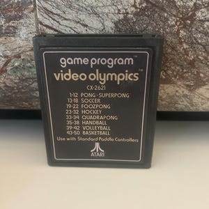 Puede incluir: Un cartucho de videojuego Atari negro con el título "video olympics" y la lista de juegos impresa en la etiqueta. La etiqueta también incluye el número de programa de juego "CX-2621" y el logotipo de Atari.