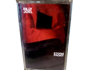 May include: A cassette tape with a red and black cover. The cassette tape is titled "Storm Front" by Billy Joel.