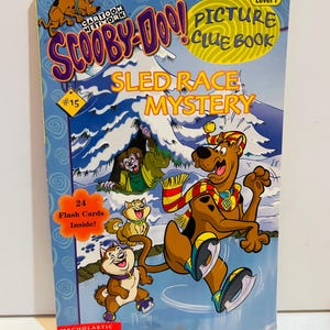 Puede incluir: Un libro de pistas ilustradas "Sled Race Mystery" de Scooby-Doo! con una portada azul. La portada presenta a Scooby-Doo patinando sobre hielo, con otros personajes de dibujos animados. El libro incluye 24 tarjetas didácticas. El logotipo de Cartoon Network es visible.