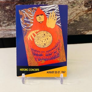 Puede incluir: Un cartel de concierto vintage para el Monterey International Pop Festival de 1967. El cartel presenta un diseño psicodélico con una figura con una túnica roja sosteniendo un globo terráqueo. El texto en el cartel dice "Monterey International Pop Festival" y "August 22-27, 1967".