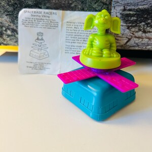 May include: A vintage Spacebase Racers Starship Viking toy. The toy features a green alien figure on a purple and pink platform, atop a blue base. The packaging includes text about the toy and its origins.
