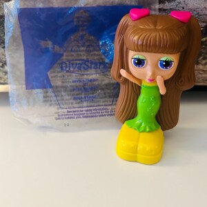 Peut inclure: Poupée Diva Starz avec des cheveux bruns, des nœuds roses en forme de cœur et de grands yeux bleus. La poupée porte une robe verte et des chaussures jaunes. Un emballage en plastique bleu avec le texte Diva Starz est en arrière-plan.