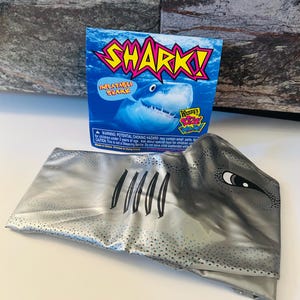 May include: An inflatable shark toy with the word "SHARK!" in large, colorful letters. The packaging features a blue ocean background and a cartoon shark. The inflatable shark is silver and gray with black details.