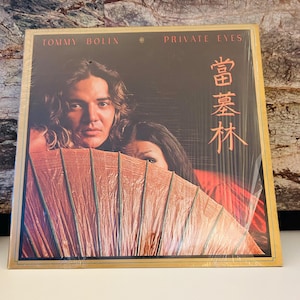 Könnte beinhalten: Ein Vinyl-Plattencover mit einem Foto von Tommy Bolin und einer Frau hinter einem großen, geöffneten Fächer. Der Albumtitel "Private Eyes" ist in Weiß auf goldenem Hintergrund gedruckt. Die japanischen Schriftzeichen "蒼葉林" sind in Rot auf der rechten Seite des Covers gedruckt.
