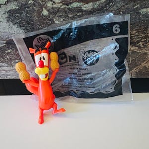 May include: An orange plastic Nutty Rodney toy from the Cartoon Network series, holding two peanut-shaped objects. The toy is in front of a clear plastic bag with the number 6 and the Cartoon Network logo.