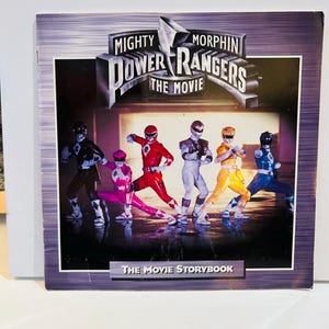 May include: A book cover for "Mighty Morphin Power Rangers: The Movie" featuring the Power Rangers in their action poses. The title is in a silver, stylized font. The background is a gradient of purple and gray. The text "The Movie Storybook" is at the bottom.