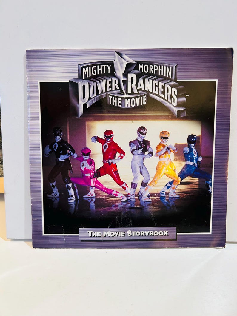 May include: A book cover for "Mighty Morphin Power Rangers: The Movie" featuring the Power Rangers in their action poses. The title is in a silver, stylized font. The background is a gradient of purple and gray. The text "The Movie Storybook" is at the bottom.