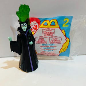 Puede incluir: Una figura de Maléfica de La Bella Durmiente de Disney, con un vestido negro y morado, una cofia verde y un accesorio verde. La figura está frente a una bolsa de McDonald's Happy Meal con el número 2.