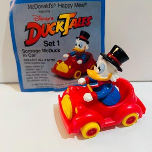 May include: A McDonald's Happy Meal toy featuring Scrooge McDuck in a red car. The toy has yellow wheels and a black top hat. The packaging reads "DuckTales" and "Set 1".