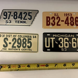 May include: Four vintage license plates from different states. The top left plate is silver with black text and reads "97-8425 53 TENN.". The top right plate is beige with red text and reads "ILL 1953 B32-486". The bottom left plate is beige with green text and reads "3-31 DIST. OF COLUMBIA $4 S-2985 THE NATION'S CAPITAL". The bottom right plate is black with white text and reads "MICHIGAN UT-36-66 53".