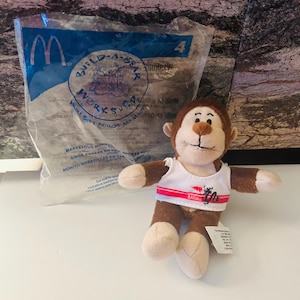 Pode incluir: Um macaco de pelúcia marrom vestindo uma regata branca com listras vermelhas e o texto "BABW". O macaco tem mãos, pés e rosto bege. Um saco plástico transparente com o texto "BUILD-A-BEAR WORKSHOP" está ao fundo.
