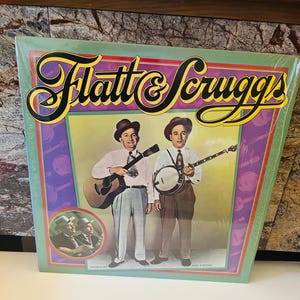 Può includere: Una copertina di un album in vinile vintage con un'illustrazione colorata del duo musicale Flatt & Scruggs. La copertina presenta i membri della band con i loro abiti caratteristici, che tengono i loro strumenti, una chitarra e un banjo. Il titolo dell'album è mostrato in modo prominente in un carattere giallo in grassetto.