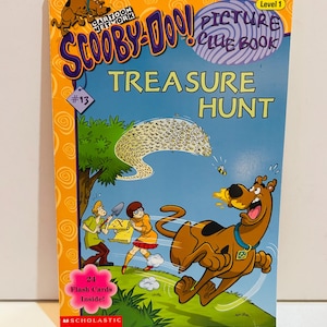 Puede incluir: Un libro de pistas ilustradas de Scooby-Doo! Treasure Hunt con una portada azul y naranja. La portada presenta a los personajes de dibujos animados Scooby-Doo, Shaggy y Velma. El libro incluye 24 tarjetas flash en el interior.