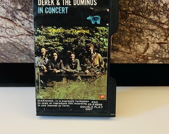May include: A black cassette tape with a photo of Derek and the Dominos on the front. The text on the tape reads "Derek & The Dominos In Concert" and "RSO CS-2-8800 Double Play 0997".