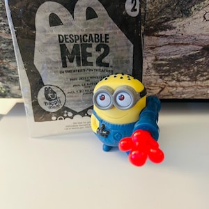 May include: A yellow Minion toy from the film Despicable Me 2, with black eyes, blue overalls, and a red hand. The toy is next to a Happy Meal bag with movie branding.