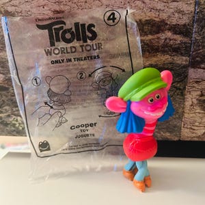 Puede incluir: Un juguete de DreamWorks Trolls World Tour, Cooper, en una bolsa de plástico transparente. El juguete es rosa con pelo azul, un gorro verde y un cuerpo rojo. La bolsa tiene el título de la película e instrucciones.