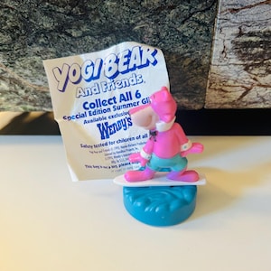 May include: A pink Yogi Bear figurine on a surfboard, part of a Wendy's "Yogi Bear and Friends" promotion. The figurine is on a blue base, with a white surfboard and a paper tag in the background.